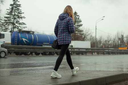The girl walks near the road. A pedestrian road along a busy highway. Girl in white shoes.