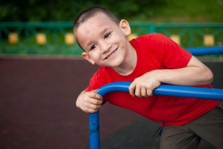 Child on merry-go-round. Boy spins on carousel for children on playground. Junior schoolboy plays alone. Child in red T-shirt with short haircut. Guy is holding on tight to handrail.の写真素材