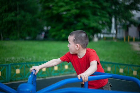 Child on merry-go-round. Boy spins on carousel for children on playground. Junior schoolboy plays alone. Child in red T-shirt with short haircut. Guy is holding on tight to handrail.の写真素材