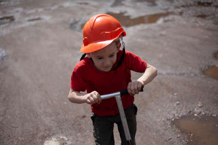 Child in orange helmet on scooter. Boy on street in summer. Child in helmet rides on dirt road. Rest of my son in summer on vacation. Younger schoolboy plays alone in the yard.の写真素材