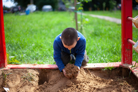 child plays in sandbox. Boy in blue jacket on playground. Preschooler touches sand with his hands. Walk in summer in the yard. Child plays alone.の写真素材