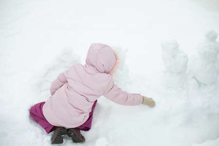Child sculpts snowman. Girl plays in snow. Baby in winter on street. Preschooler in warm clothes plays alone.の写真素材