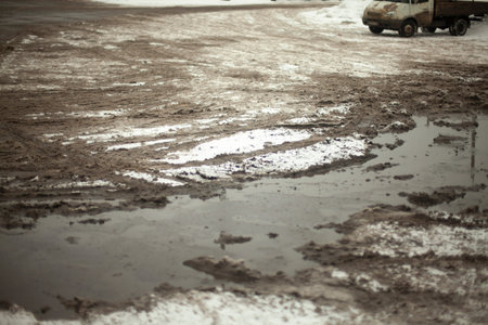 Muddy puddle of melted snow on road. Uncleaned parking. Asphalt is covered with layer of snow.