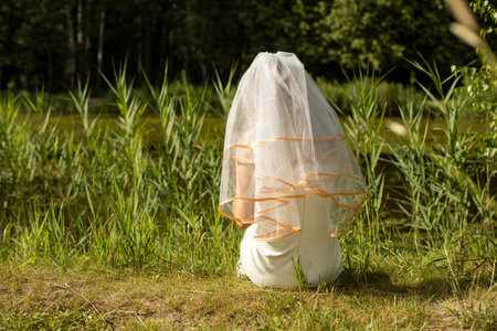 Bride on the shore of the lake. Girl in wedding dress in park. White dress girls. Veil on head. Outfit details.の写真素材