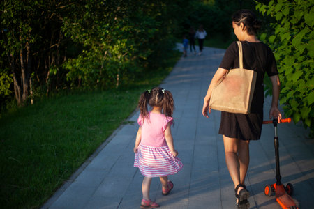 Mother with child walks through the park. Walking with child in summer. Asian family on street. Mother carries her daughter's scooter.の写真素材
