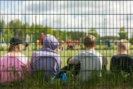 Children in the stadium. Teenagers on sports field. Schoolchildren sit on grass behind net. Fence around football field. Spectators at sports competition.の写真素材