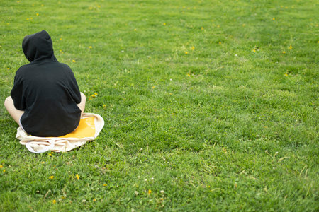 Teenager in black jacket in park. Girl in black hood sits on green grass. alienated man. Teenage crisis.の写真素材