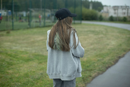 girl in the park. Teenager walks on street. Black cap and long hair. Gray jacket. girl in summer.の写真素材