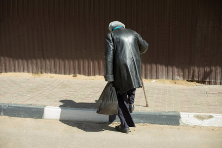 Old woman with walking stick walks down road. Pensioner in Russia. black coat. Elderly man walks along the fence. Old age city.の写真素材