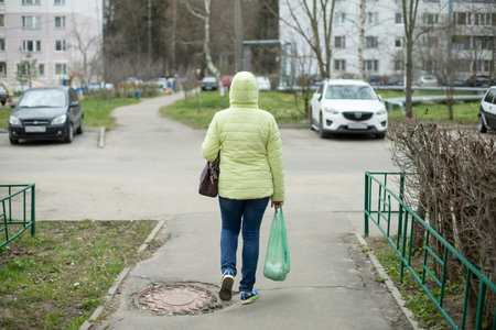 Woman walks with bag in her hand down street. Girl in green jacket. Person walks out of store with groceries in bag.の写真素材
