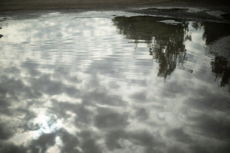 Clouds are reflected in puddle. Cloudy weather outside. Huge puddle on asphalt. Surface of water.の写真素材