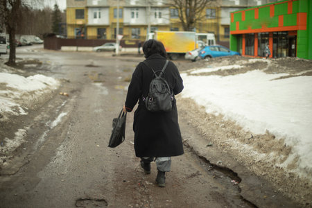 Woman walks along dirt road in city. Man in black jacket with backpack. Woman in Eastern Europe.の写真素材
