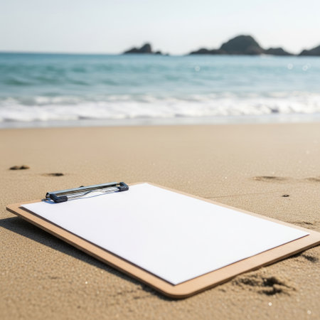 A clipboard with a blank sheet of paper lies on the sandy beach near the shoreline with gentle waves and rocky formations in the background.の素材
