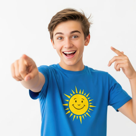 A happy young boy, likely a teenager, points directly at the viewer while smiling broadly, wearing a blue t-shirt with a smiling sun graphic against a white background.の素材
