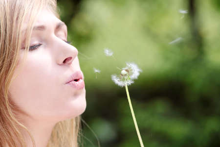 The blonde sits in park on a grass with a dandelion in hands and blowingの写真素材