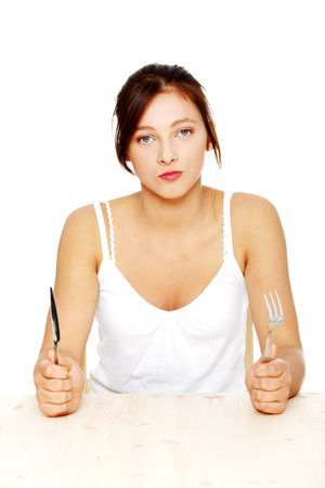 Hungry pretty caucasian teenage girl sitting behind the table with fork and knife. Isolated on...