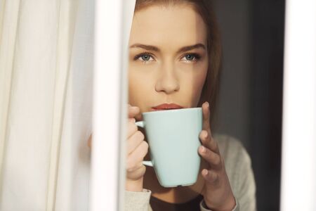 Beautiful caucasian woman drinking hot coffee or tea and looking through window. Indoor background.の写真素材