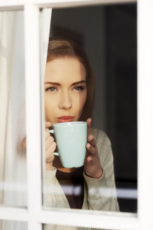 Beautiful caucasian woman drinking hot coffee or tea and looking through window. Indoor background.の写真素材