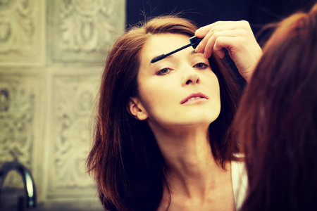 Woman using mascara in bathroom mirror.