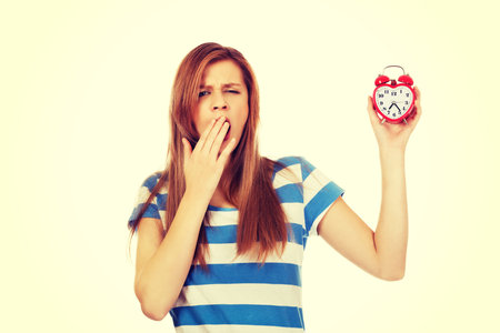 Yawning teenage woman holding alarm clock.