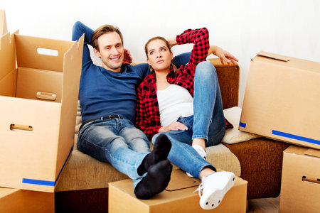 Happy tired couple sitting on couch in new home with cordboard boxes around.