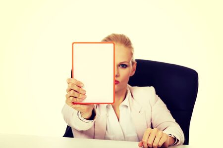 Serious business woman sitting behind the desk and holding board with ban.の写真素材