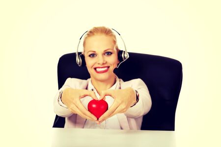 Happy call center woman sitting behind the desk and holding heart model.の写真素材