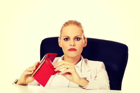 Worried business woman sitting behind the desk with empty wallet.