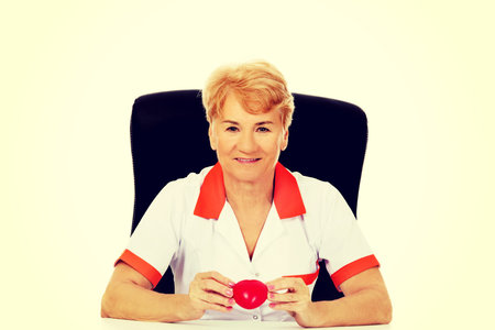 Smile elderly female doctor or nurse sitting behind the desk and holds heart toy.