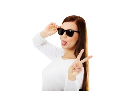 Happy brunette woman in sunglasses shows tongue and victory sign.の写真素材