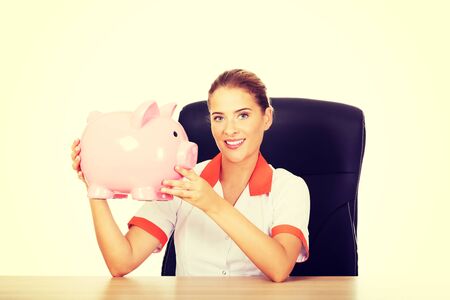 Young smile female doctor or nurse sitting behind the desk and holding a piggybank.の写真素材