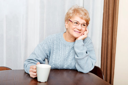 Senior woman sitting at the desk and drinking coffee or tea.