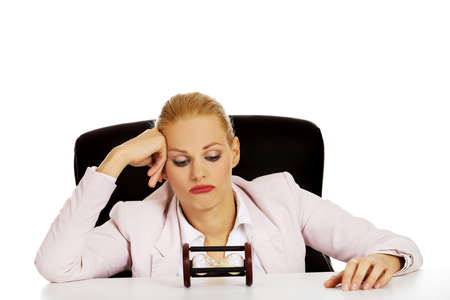 Business woman sitting behind the desk and looking for sandglass.の写真素材