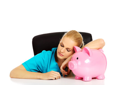 Young tired female doctor or nurse sitting behind the desk and holding a piggybank.の写真素材