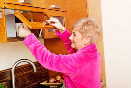 Smile senior woman taking mug from a kitchen cabinet.の写真素材