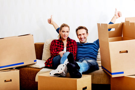 Happy tired couple sitting on couch in new home with cordboard boxes around