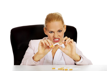 Angry business woman sitting behind the desk and breaking down cigaretteの写真素材