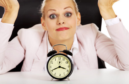 Surprised business woman lsitting behind the desk with alarm clockの写真素材