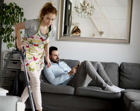 Woman doing chores using vacuum cleaner on carpet and lazy man on sofaの写真素材