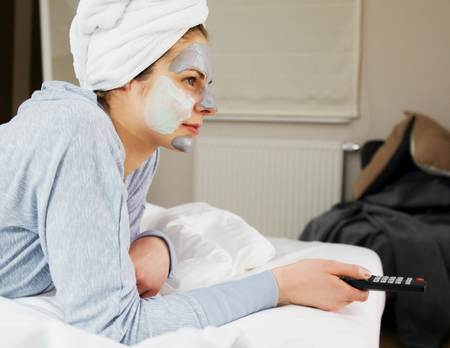 Young woman with clay mask on face watching tv at home.の写真素材
