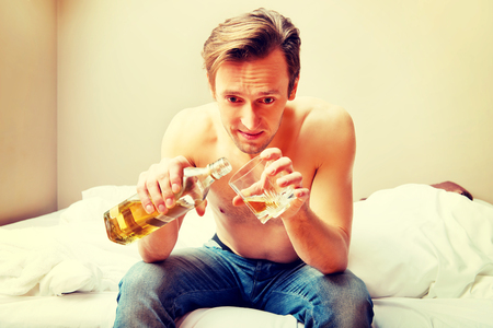Drunk man sitting on bed and drinking whiskeyの写真素材