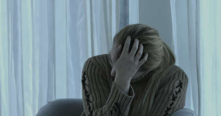 Depressed sad woman at home sitting on the couch, looking down and touching her forehead, loneliness and pain conceptの写真素材