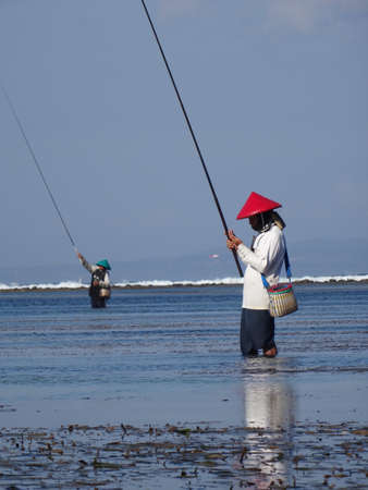 Typical Balinese fisherman is standing in the shallow water while checking his bite of the fishing rod at Sanur Beach in Bali, Indonesiaのeditorial素材