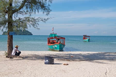 Otres Beach, Sihanoukville, Cambodia - 30th December 2018 : Beautiful seashore of Otres beach with a boatman sitting on the white sand and two typical long-tale boats in the backgroundのeditorial素材