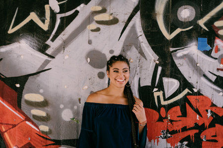 Young smiling African American woman leaning on a graffiti wall, beautiful girl, urban portrait. Summer in the city. Millenial. Authentic, unposed. Black beauty. Dark skin. Braids. Youth culture.のeditorial素材