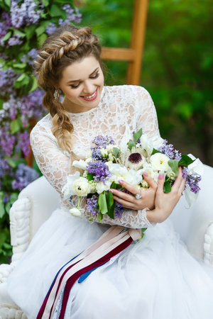 A smiling bride is sitting in a chair and holding a bridal bouquet.の写真素材