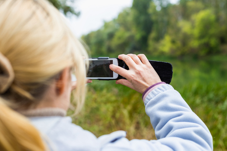 A blonde woman using a phone takes a photo of a beautiful landscape.の写真素材
