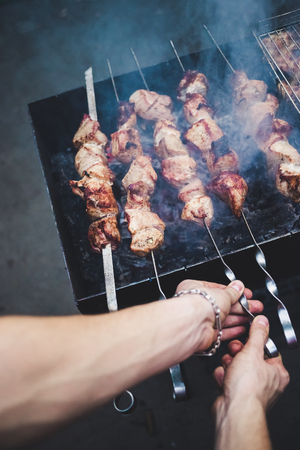 Grilled kebab cooking on metal skewers (grill). Roasted meat cooked at barbecue with smoke. Close up BBQ fresh pork meat chop slices. Traditional eastern dish, shish kebab. Grill on charcoal and flame, picnic, street food. Toned, style, classic photo.の写真素材