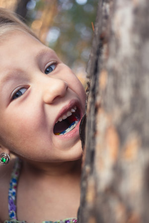 A little girl with a wobbly baby tooth in her mouth is trying to bite a tree trunk