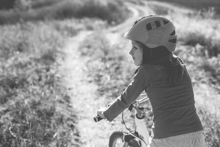 Little girl on a bike wearing a helmet on a dirt road in the field. Old monochrome photo.の写真素材
